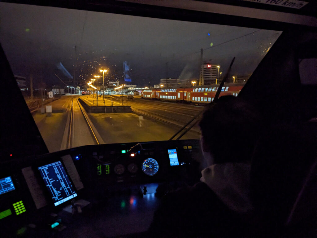 David im Führerstand kurz vorm Kasseler Hbf. Von hinten fotografiert. Man sieht aus der Frontscheibe einen gelb beleuchteten Bahnsteig bei Nacht.