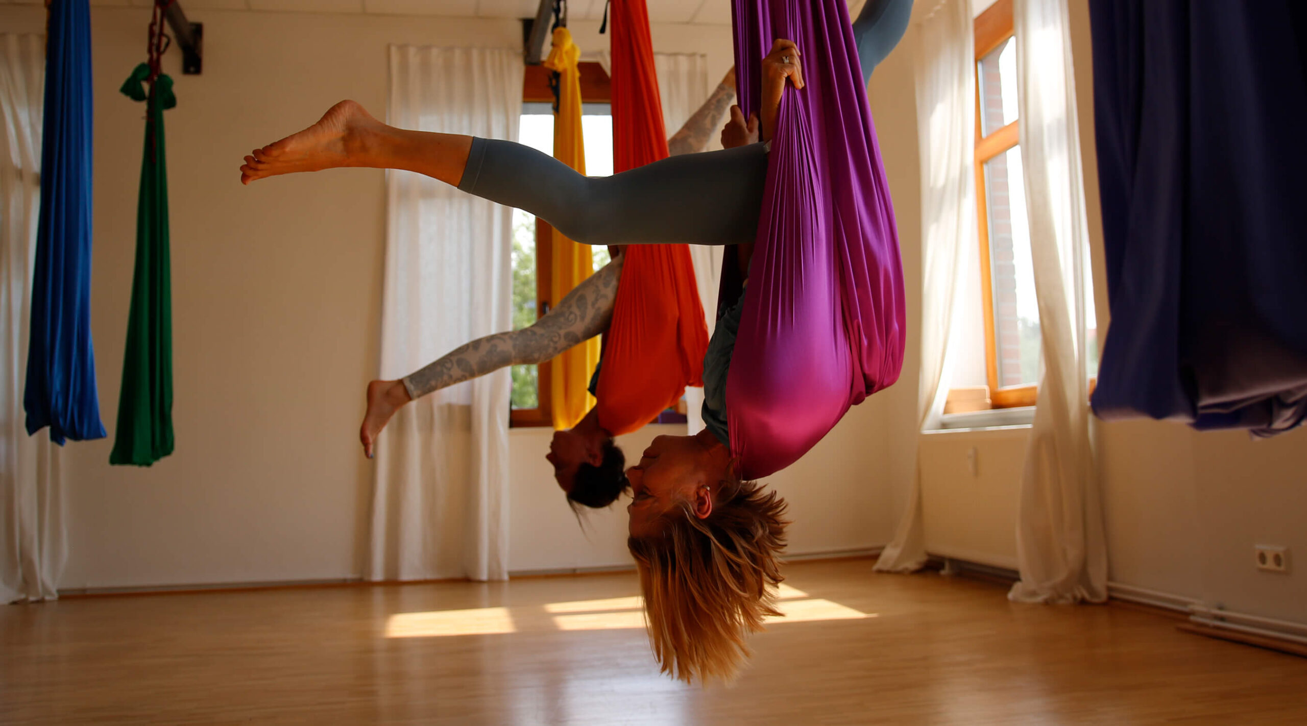 Zwei Frauen hängen kopfüber mit Spagat in Aerial Yoga-Tüchern von der Decke. | Foto: Maarten Hoffmeyer